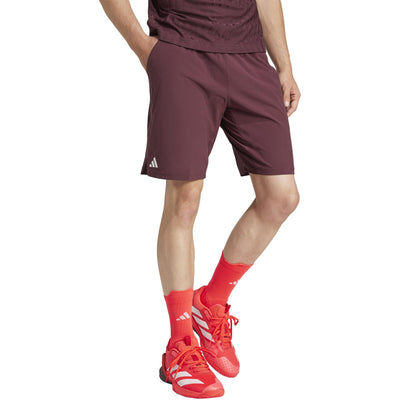 A person wears adidas Ergo Men's Tennis Shorts in ruby, crafted from recycled materials. Their matching t-shirt complements vibrant red socks and athletic shoes with white stripes and the iconic adidas logo against a plain white backdrop.