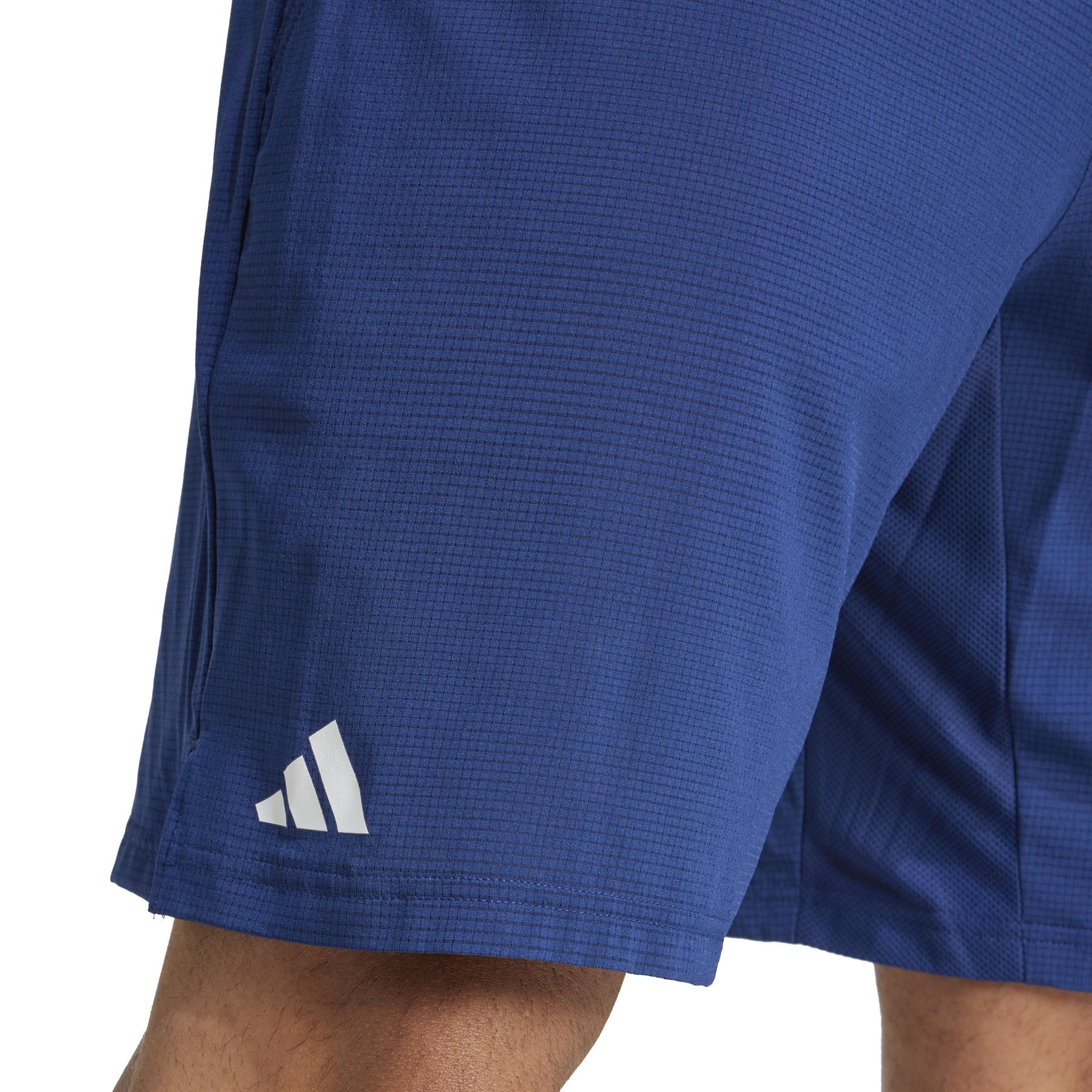 A person stands wearing ADIDAS Ergo Men's Tennis Shorts in navy, showcasing the adidas logo on the lower left. These shorts are lightweight with a subtle textured pattern and crafted from recycled materials.