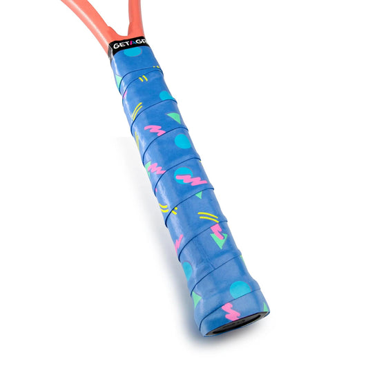 A close-up of a tennis racket handle wrapped with the Get A Grip Cutting Shapes Tennis Overgrip in blue. The premium, non-slip material showcases vibrant designs, featuring pink zigzags and green circles. The frame is partially visible at the top, highlighting the eye-catching overgrip by Get A Grip.