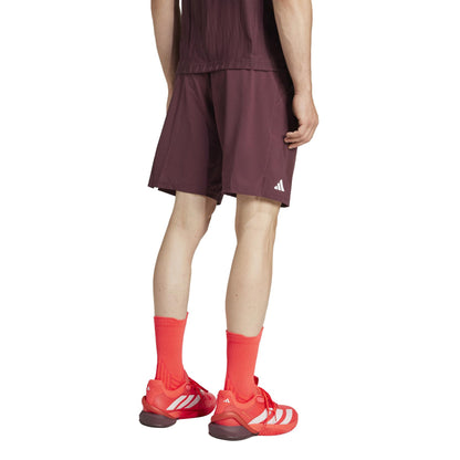 A person dressed in a dark maroon athletic outfit with bright red socks and sneakers stands on a white background. They wear ADIDAS Ergo Men's Tennis Shorts in Ruby, crafted from recycled materials for an eco-friendly choice.
.