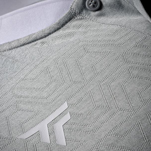 Close-up of a silver textured polyester mesh fabric with a geometric pattern. A small button is visible at the top, and there's a white logo with overlapping "T" shapes near the bottom, indicative of the breathable design of the Tecnifibre Men's Team Tennis Mesh Polo Shirt.