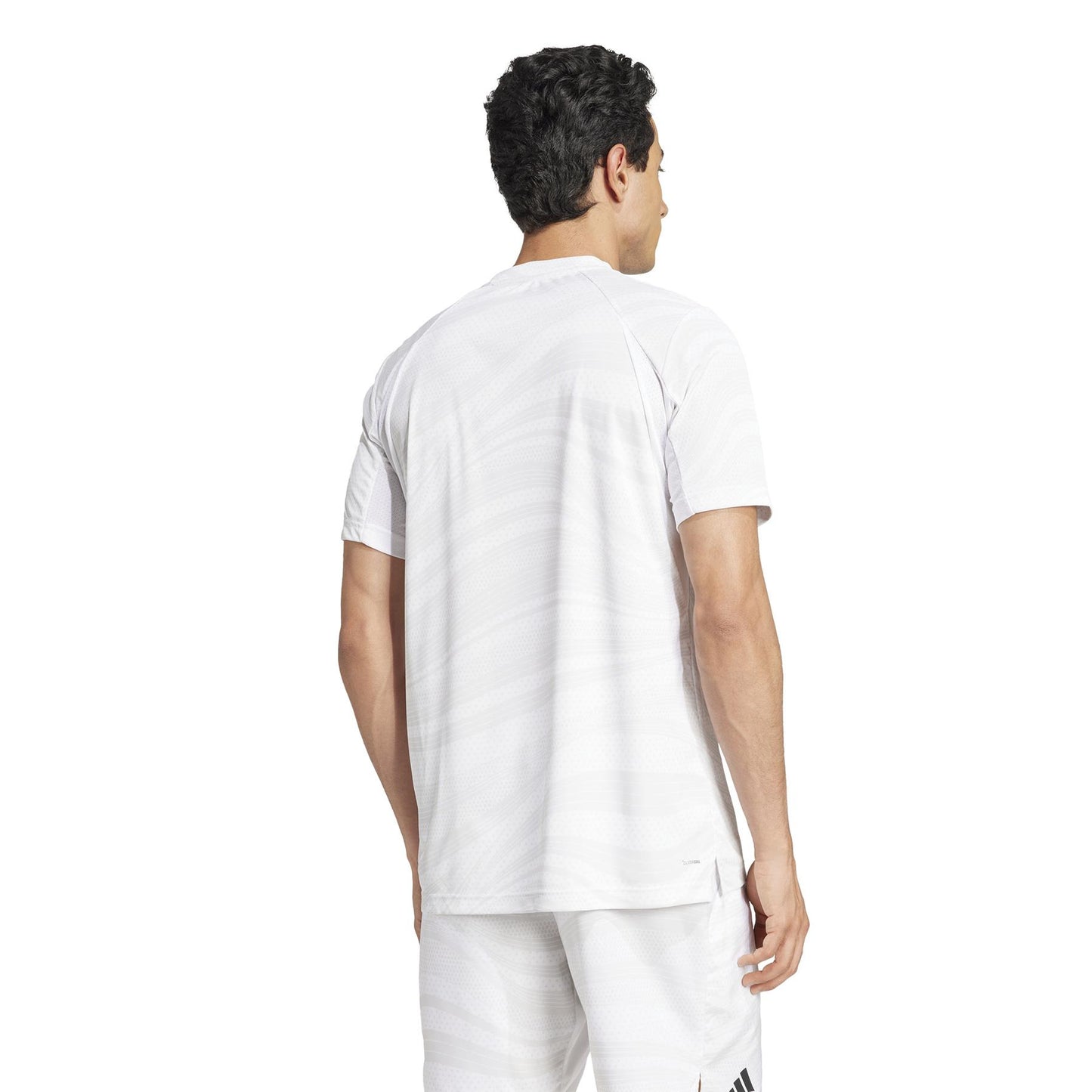 A person with short, dark hair, facing away from the camera, is wearing an ADIDAS Men's Club Graphic Tennis T-Shirt in White/Grey made of recycled materials. The jersey and shorts use AEROREADY technology and subtle designs for a sleek look.