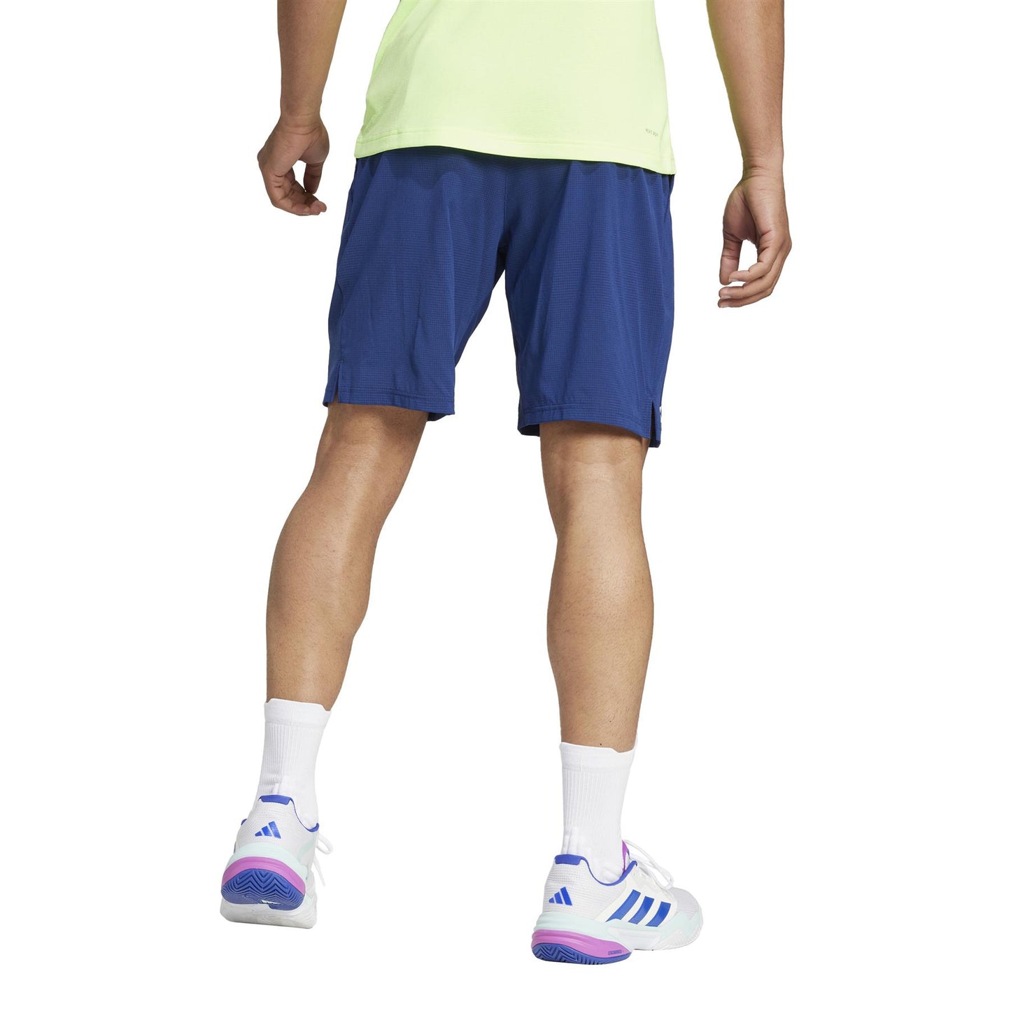 Wearing a lime green shirt and ADIDAS Ergo Men's Tennis Shorts in navy, a person stands on a white background. The look is completed with white socks and sneakers featuring vibrant blue and pink accents.