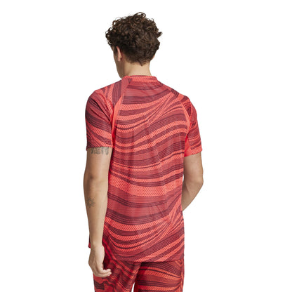 A person with curly hair wears an ADIDAS Mens Club Graphic Tennis T-Shirt in red with a black abstract design, made from recycled materials. The wavy pattern extends to their shorts. They have a small tattoo on their upper arm and stand against a plain white background.