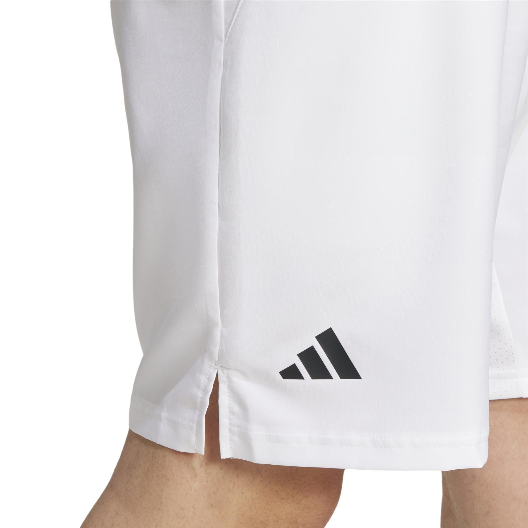 Close-up of someone wearing ADIDAS Men's 3 Stripe Tennis Shorts in white, showcasing the black logo near the hem. The lightweight fabric uses AEROREADY technology and has a small side slit to enhance movement.