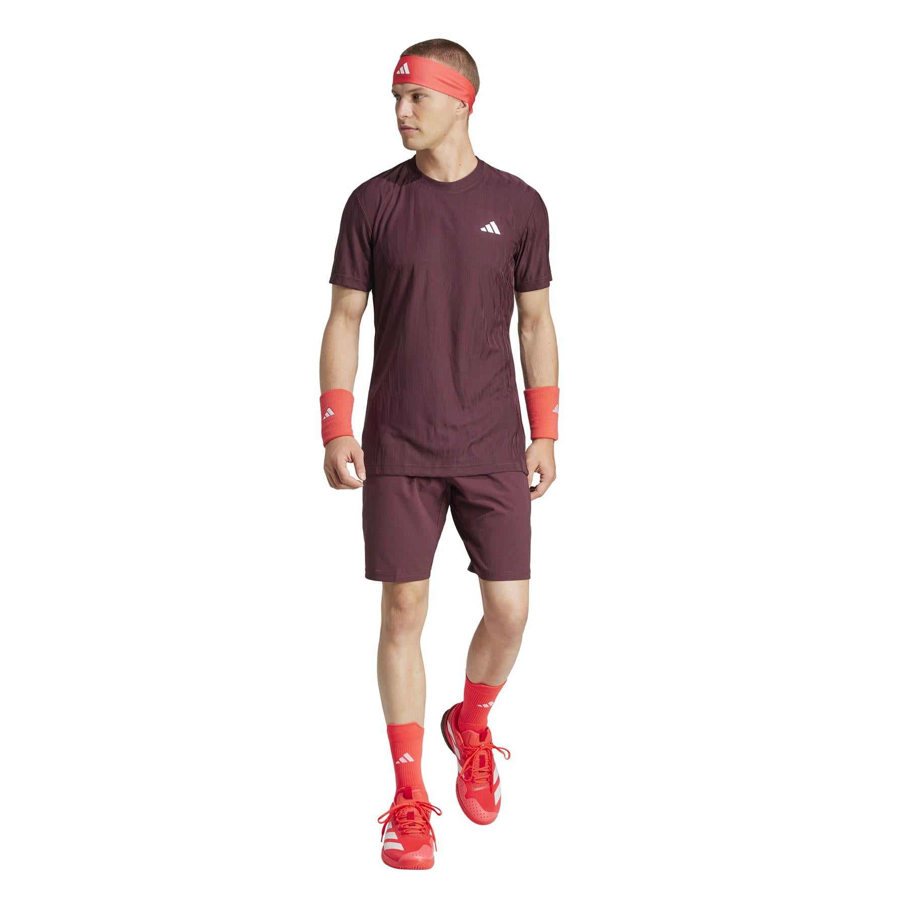 A person in a maroon athletic outfit, including ADIDAS Ergo Men's Tennis Shorts in Ruby, adorned with red accessories—headband, wristbands, shoes, and socks—poses against a white background while looking to the side.