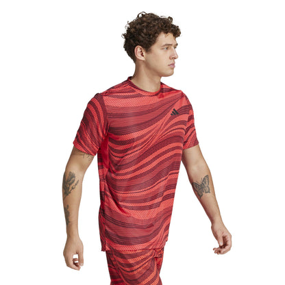 A person with curly hair is walking while wearing a red, wavy-patterned athletic shirt and matching shorts. Their Adidas Men's Club Graphic Tennis T-Shirt in red, made from recycled materials with AEROREADY technology, sports a small chest logo. Tattoos adorn their arms.