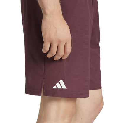 A person stands with one hand by their side, showcasing ruby ADIDAS Ergo Men's Tennis Shorts. The focus is on the white adidas logo and their recycled material construction, highlighting their lightweight design.