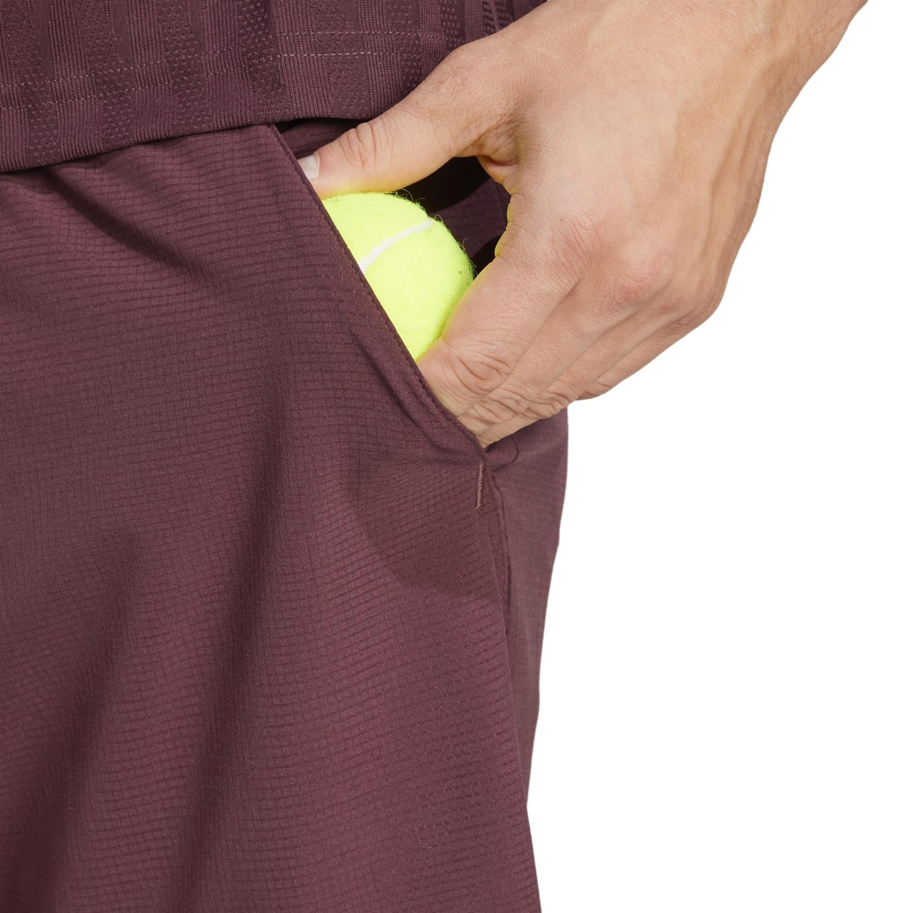 A person placing a yellow tennis ball into the side pocket of textured adidas Ergo Men's Tennis Shorts in Ruby, made from recycled materials.