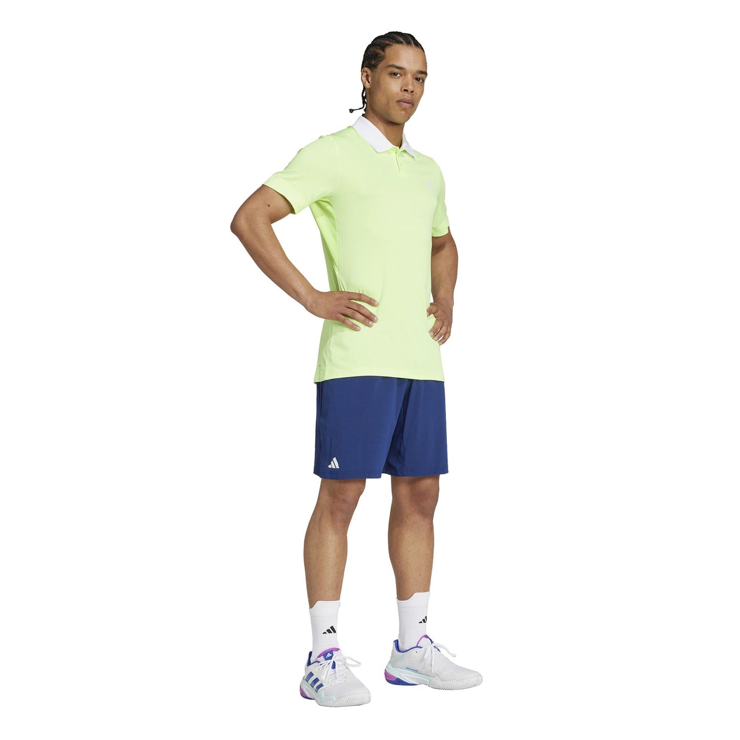 A person stands confidently with braided hair, wearing a lime green polo and ADIDAS Ergo Men's Tennis Shorts - Navy. Their sporty look is enhanced by white socks and sneakers. The eco-friendly shorts made from recycled materials pop against the plain white background.