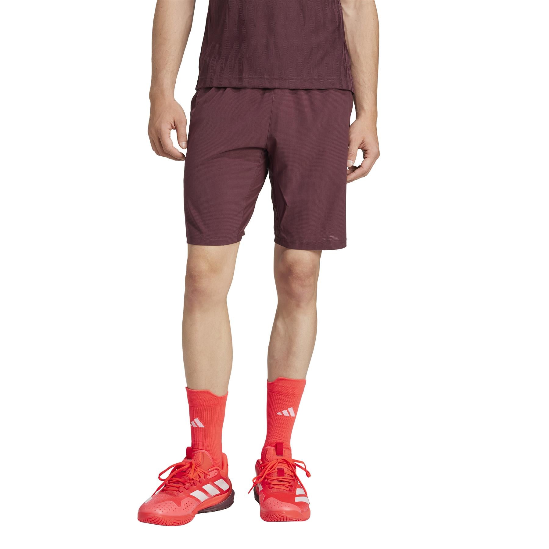 A person wears lightweight ruby shorts similar to the ADIDAS Ergo Mens Tennis Shorts by adidas, crafted from recycled materials, paired with a matching top, bright orange socks, and orange-red shoes, standing against a white background. The image is cropped at the shoulders.