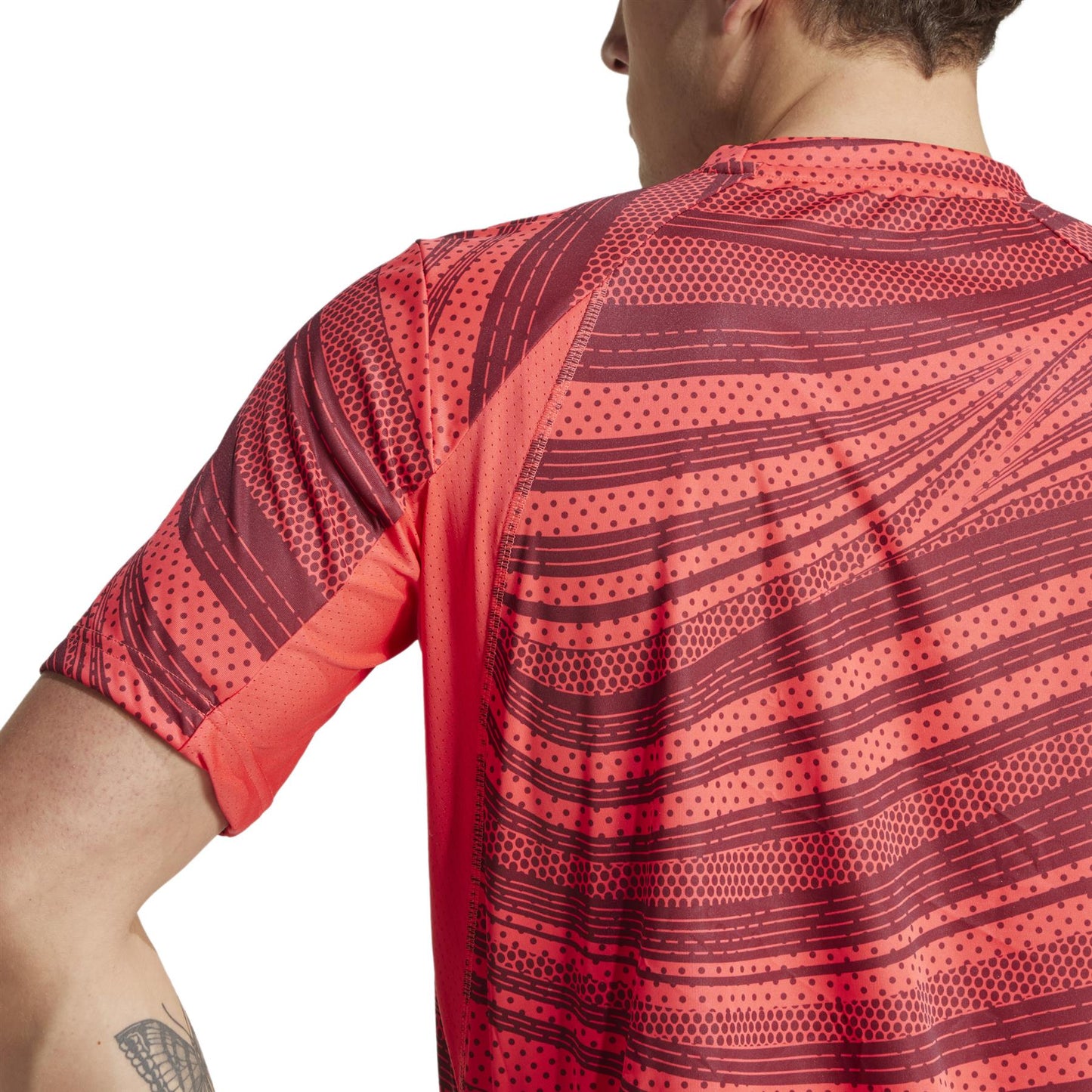 The image displays someone from behind wearing an adidas Men's Club Graphic Tennis T-Shirt in red, featuring a patterned athletic design with horizontal and diagonal lines plus small contrasting dots. The shirt incorporates recycled materials, and their right arm is bent at the elbow.