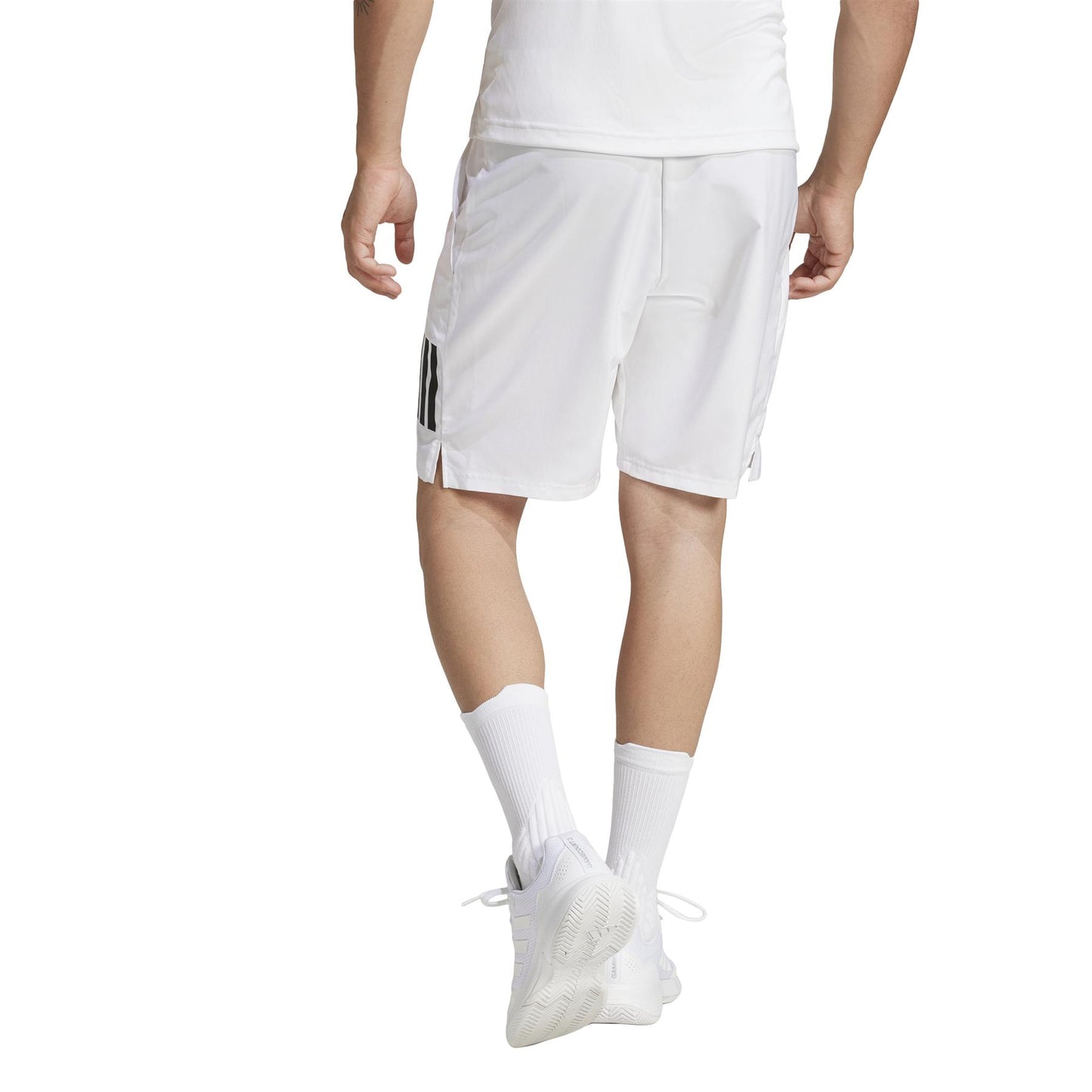 The image shows a person from behind in an athletic ensemble featuring adidas Men's 3 Stripe Tennis Shorts - White, crafted from recycled materials. Their outfit includes a plain white t-shirt, white sneakers, and simple calf-length white socks with understated style.