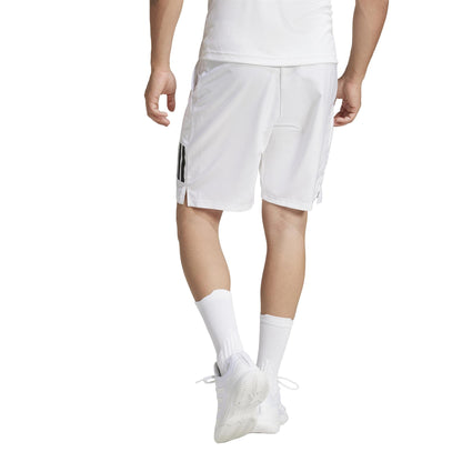 The image shows a person from behind in an athletic ensemble featuring adidas Men's 3 Stripe Tennis Shorts - White, crafted from recycled materials. Their outfit includes a plain white t-shirt, white sneakers, and simple calf-length white socks with understated style.