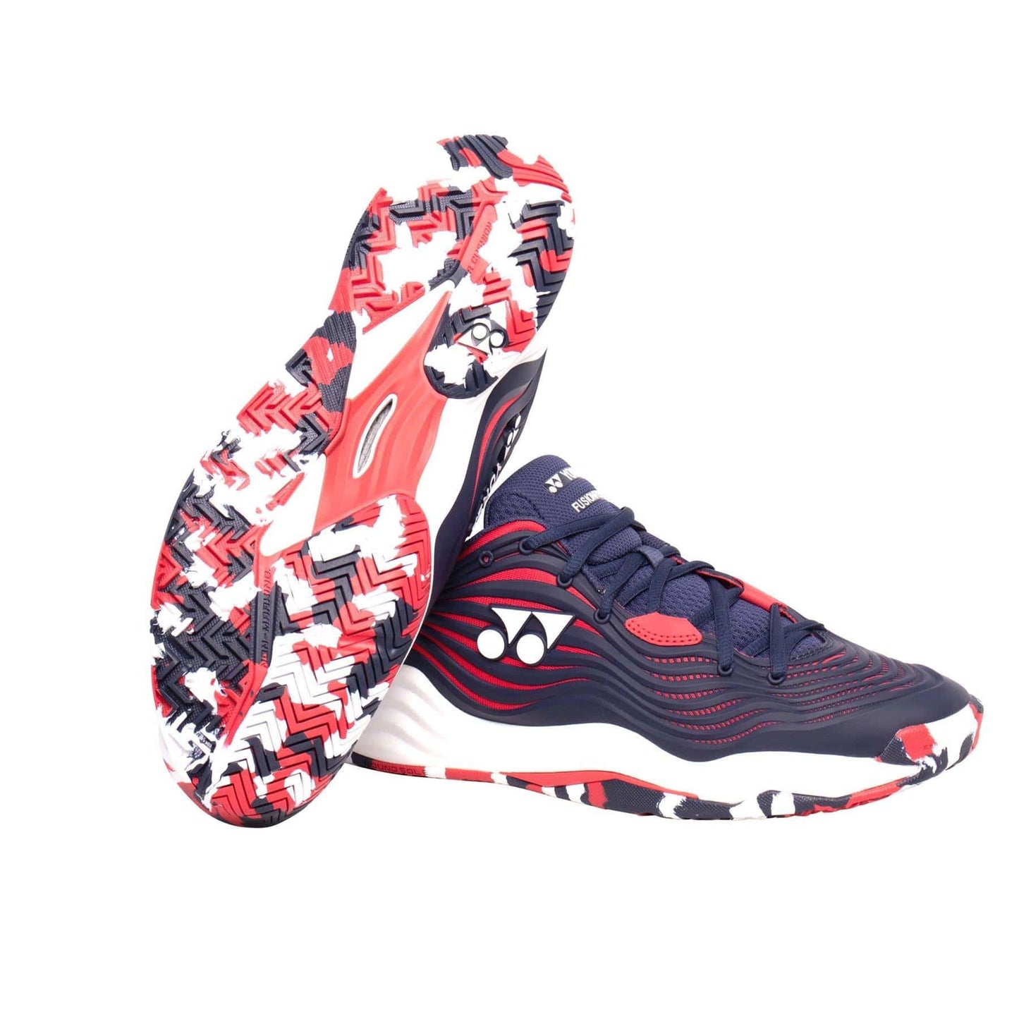 A pair of Yonex Power Cushion FusionRev 5 men's tennis shoes showcases vibrant navy and red patterns. The soles feature a striking zigzag design in shades of red, black, and white, enhanced with Power Cushion technology. The Yonex logo stands out prominently on the side. One shoe is upright while the other rests on its side.