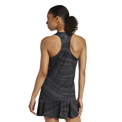 A woman showcases the ADIDAS Women's Club Graphic Tennis Tank Top in black, featuring a racerback design and pleated skirt made from recycled materials. Her ponytail complements the sleek outfit, which uses Climacool technology to keep her cool against a plain white backdrop.