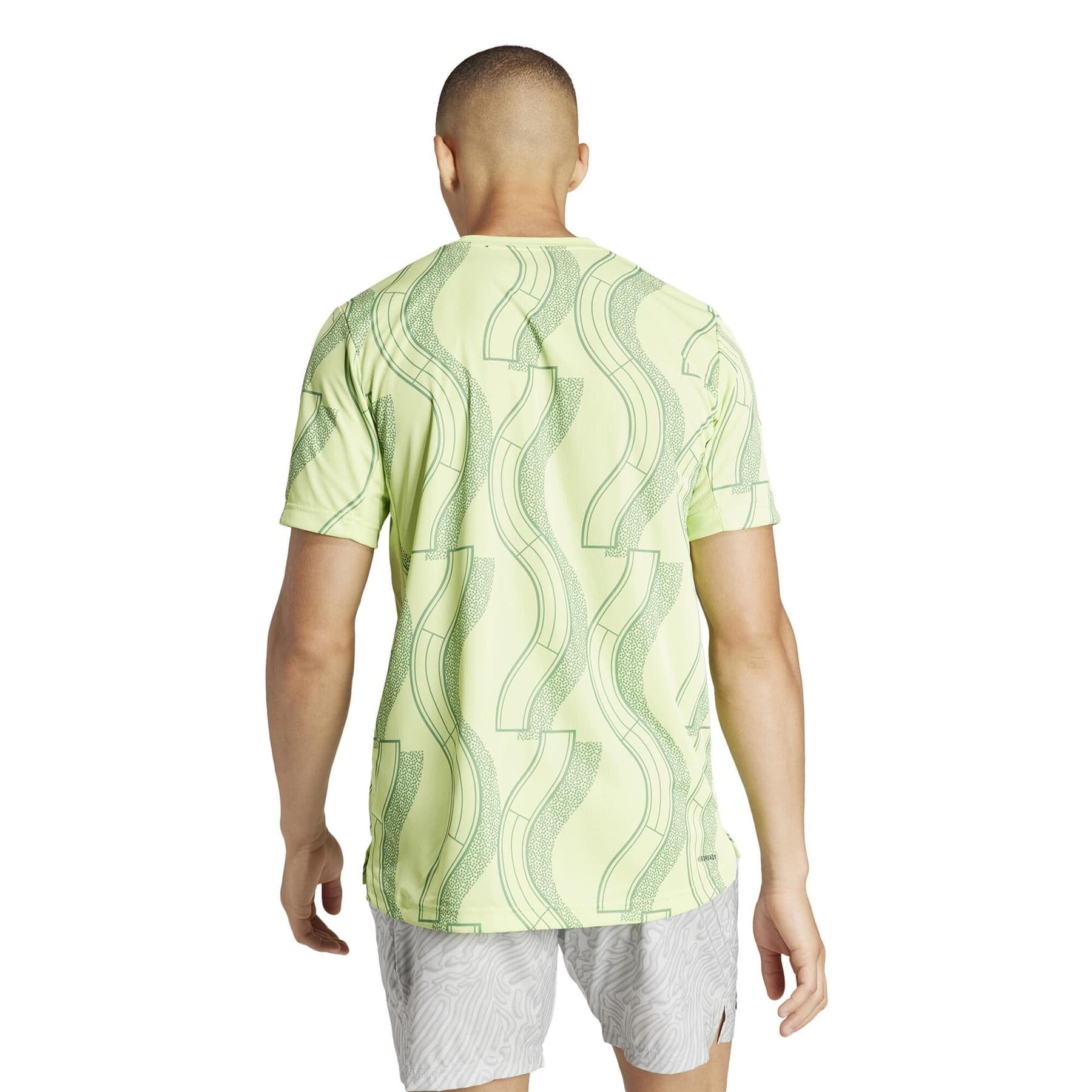 The person is wearing an ADIDAS Men's Club Graphic Tennis T-Shirt in green, featuring a wavy design, paired with light gray shorts. Made from recycled materials, the AEROREADY fabric provides both comfort and style. They are posed against a plain white background, giving the scene a modern feel.