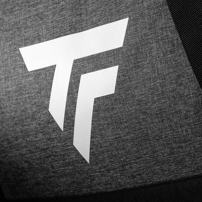 A close-up of a white, stylized "TF" logo is displayed on a gray fabric with a textured surface. The high-contrast composition highlights the sleek design of the Tecnifibre All Vision Tennis Duffle Bag - Grey, showcasing the clean lines of the Tecnifibre logo against the darker fabric.