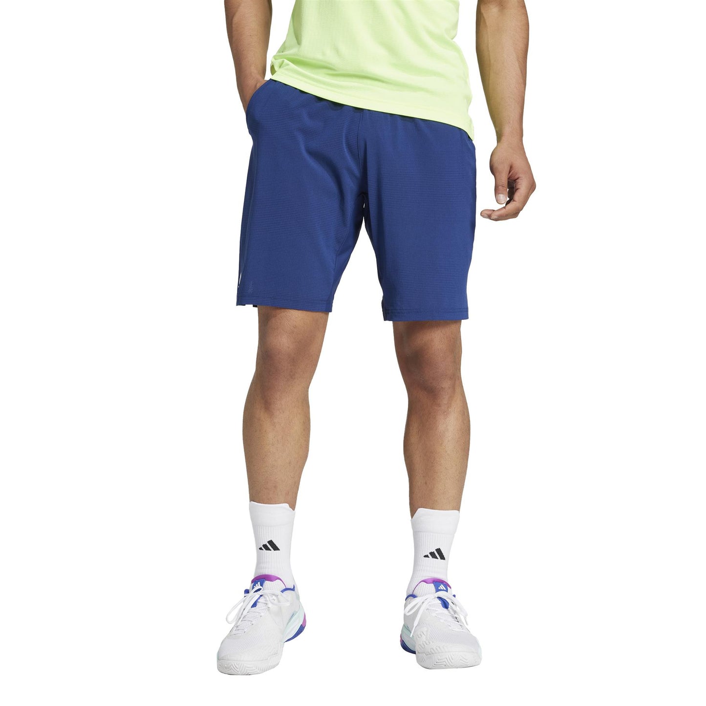 A person in a neon green shirt, adidas Ergo Men's Tennis Shorts - Navy made from recycled materials, with white socks and white athletic shoes with purple accents. The cropped photo highlights their casual sporty outfit featuring lightweight navy tennis shorts.