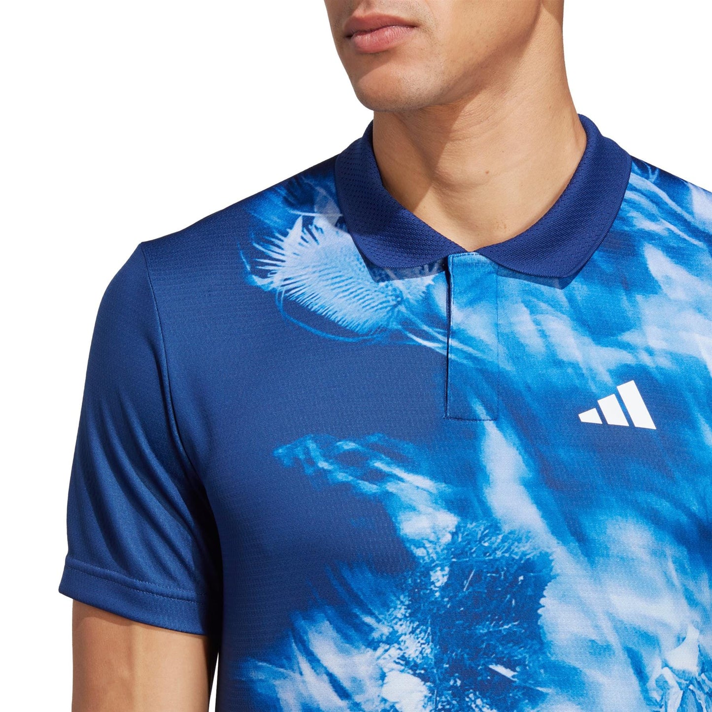 The image displays an individual in the adidas Melbourne Mens Freelift Tennis Polo Shirt - Blue by adidas. The shirt features a blue and white abstract pattern with a white logo on the front, and incorporates HEAT.RDY technology. Captured from shoulders to upper torso while facing right, the image embodies Australian Open vibes.