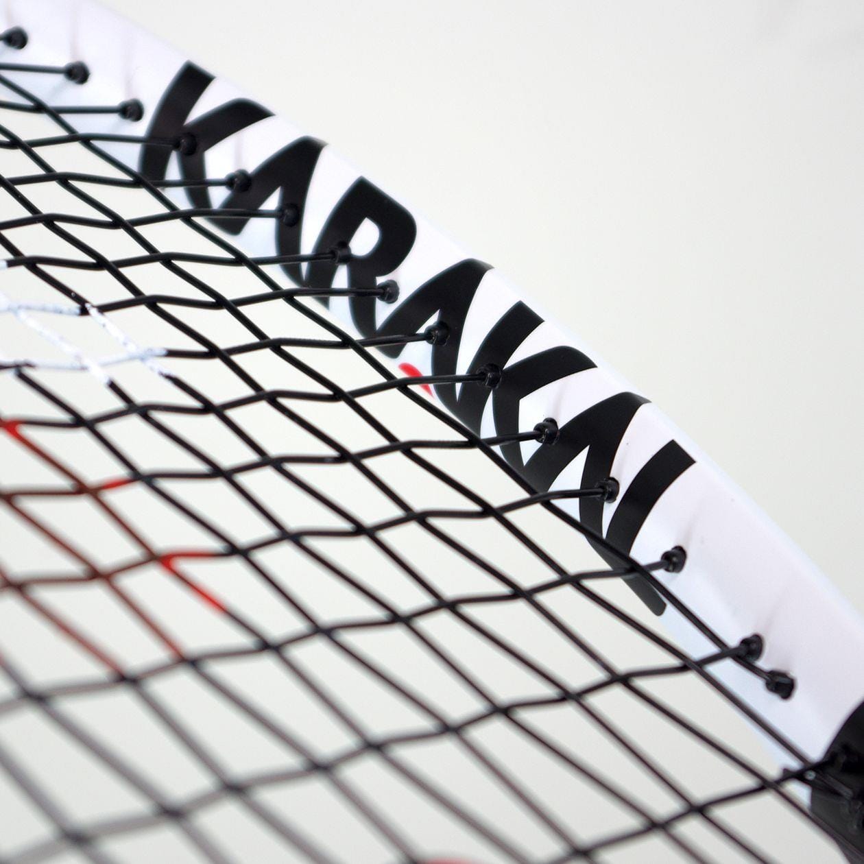 Close-up of the Karakal Comp 27 Tennis Racket's strings and frame, highlighting the brand name "Karakal" in black letters on a white 7050 Alumag Alloy frame. The image emphasizes the intricate pattern of black strings, ideal for those seeking a reliable beginner-level racket.